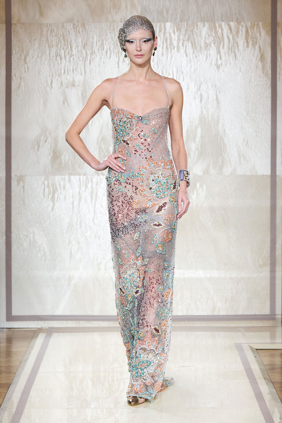 Model on a runway wearing a sheer, pastel-toned gown covered in multicolored embroidery and crystals, with a beaded headpiece.