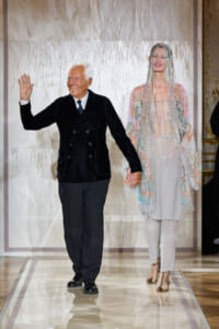 Older man in a black velvet suit waves while walking hand-in-hand with a model wearing an embellished, beaded cape and a fringe headpiece on a runway backdropped by a light-toned wall.