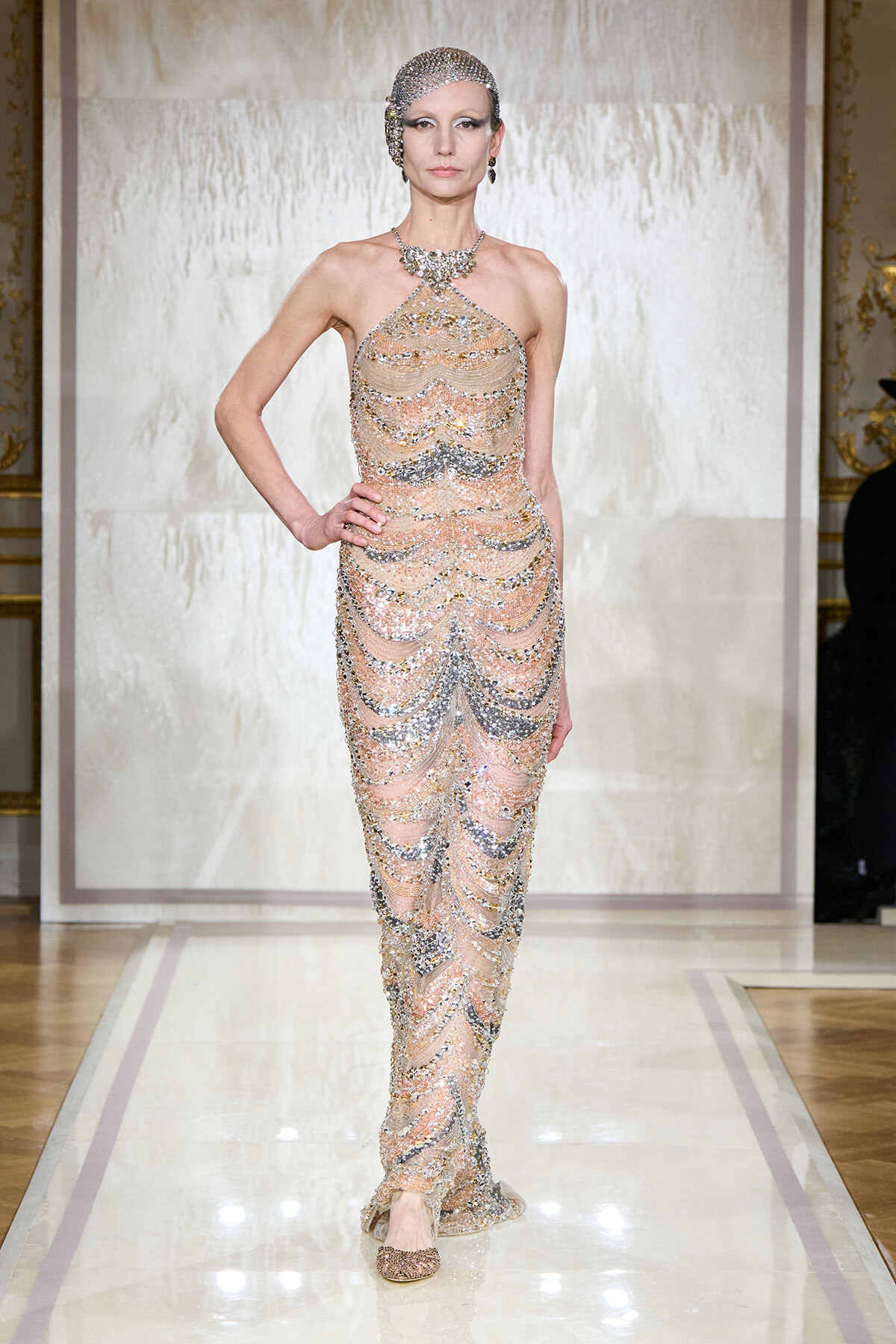 Model walks a fashion runway wearing a sheer nude gown covered in cascading silver beads and sequins, with a matching headpiece and necklace.