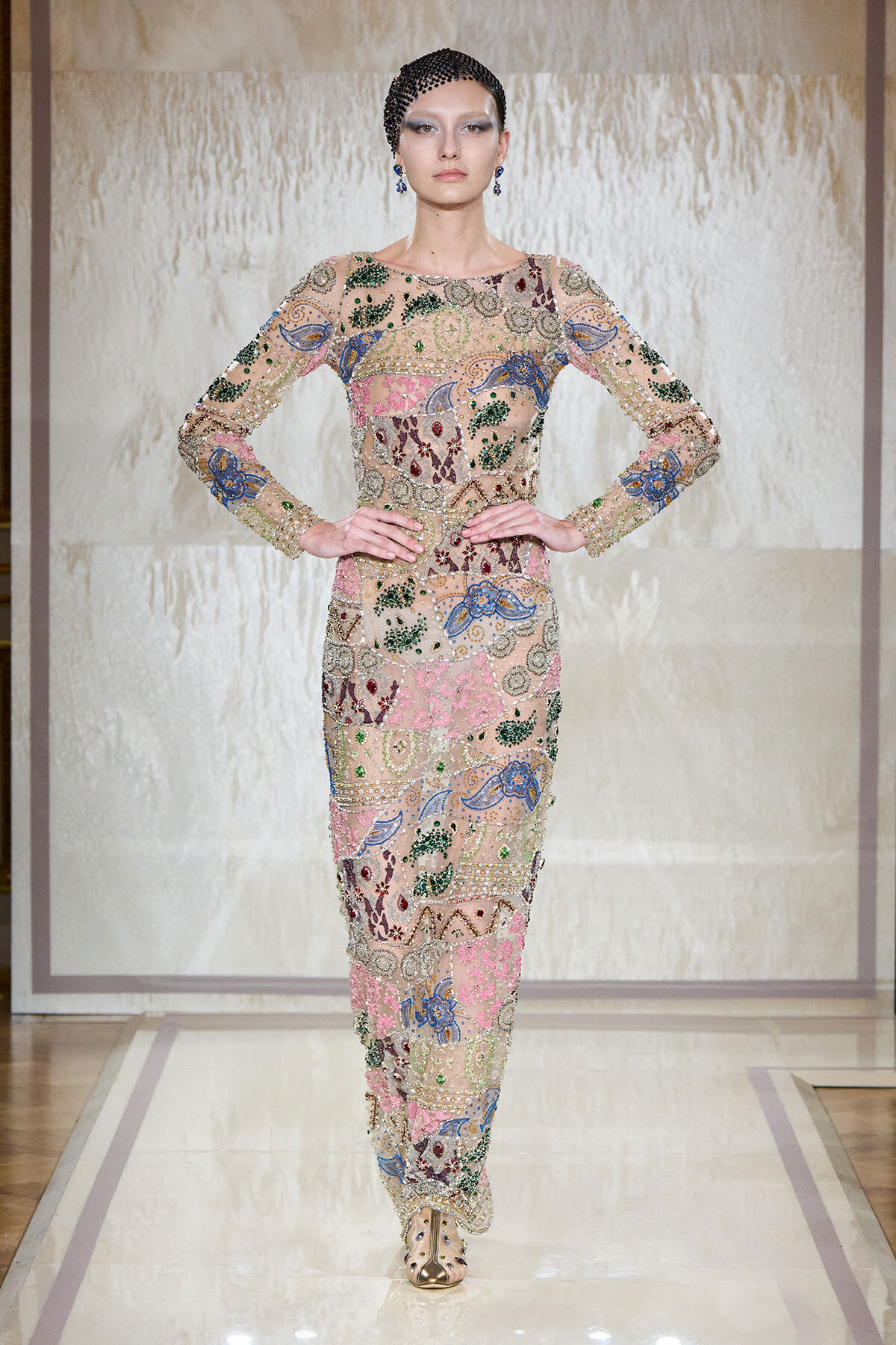 Model on a runway wearing a full-length sheer gown covered in multicolored beadwork and embroidery with long sleeves, hands on hips.