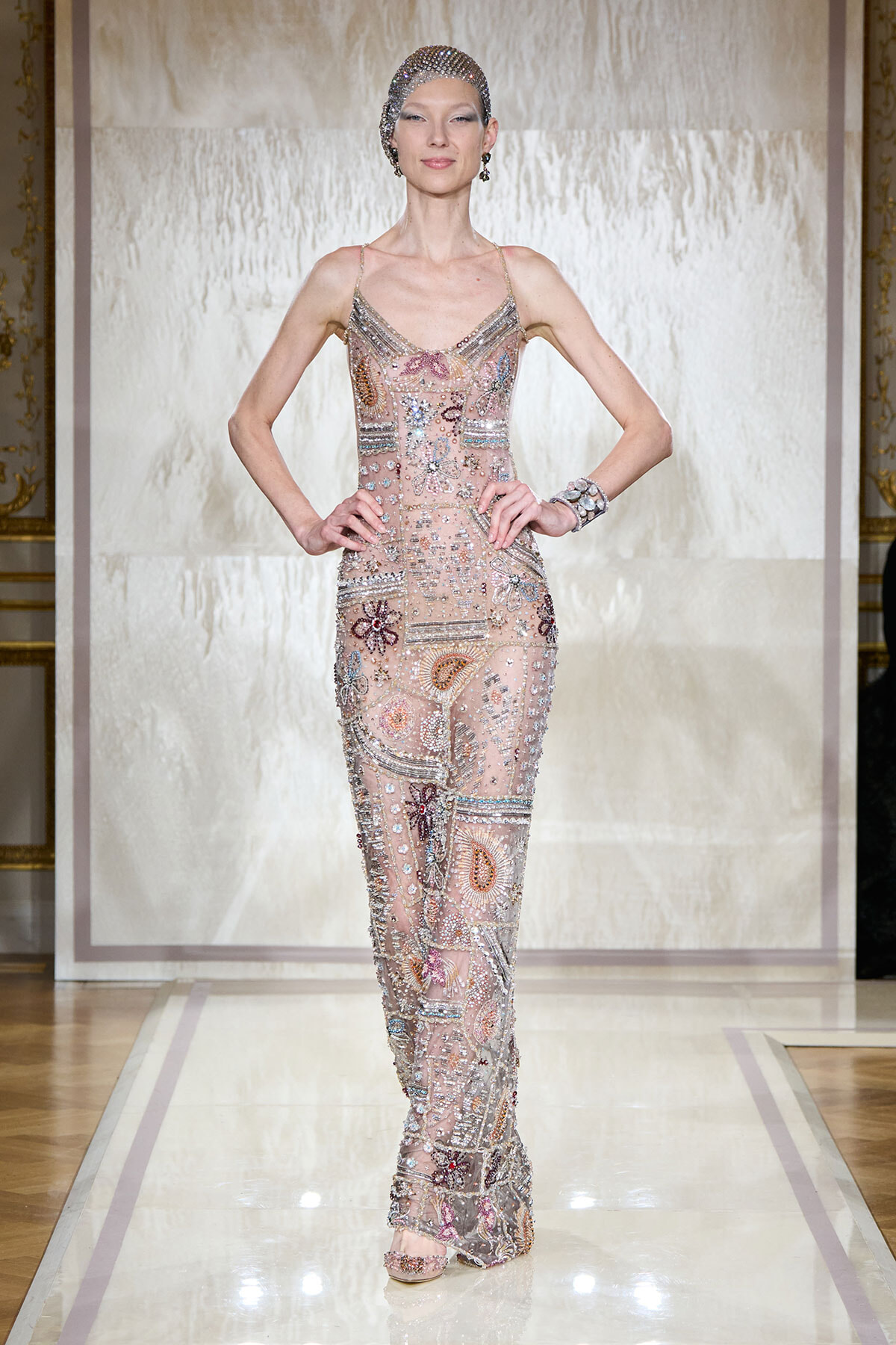 Model on a runway wearing a nude, sheer jumpsuit heavily embellished with beads and sequins, plus a matching beaded cap and jewelry.