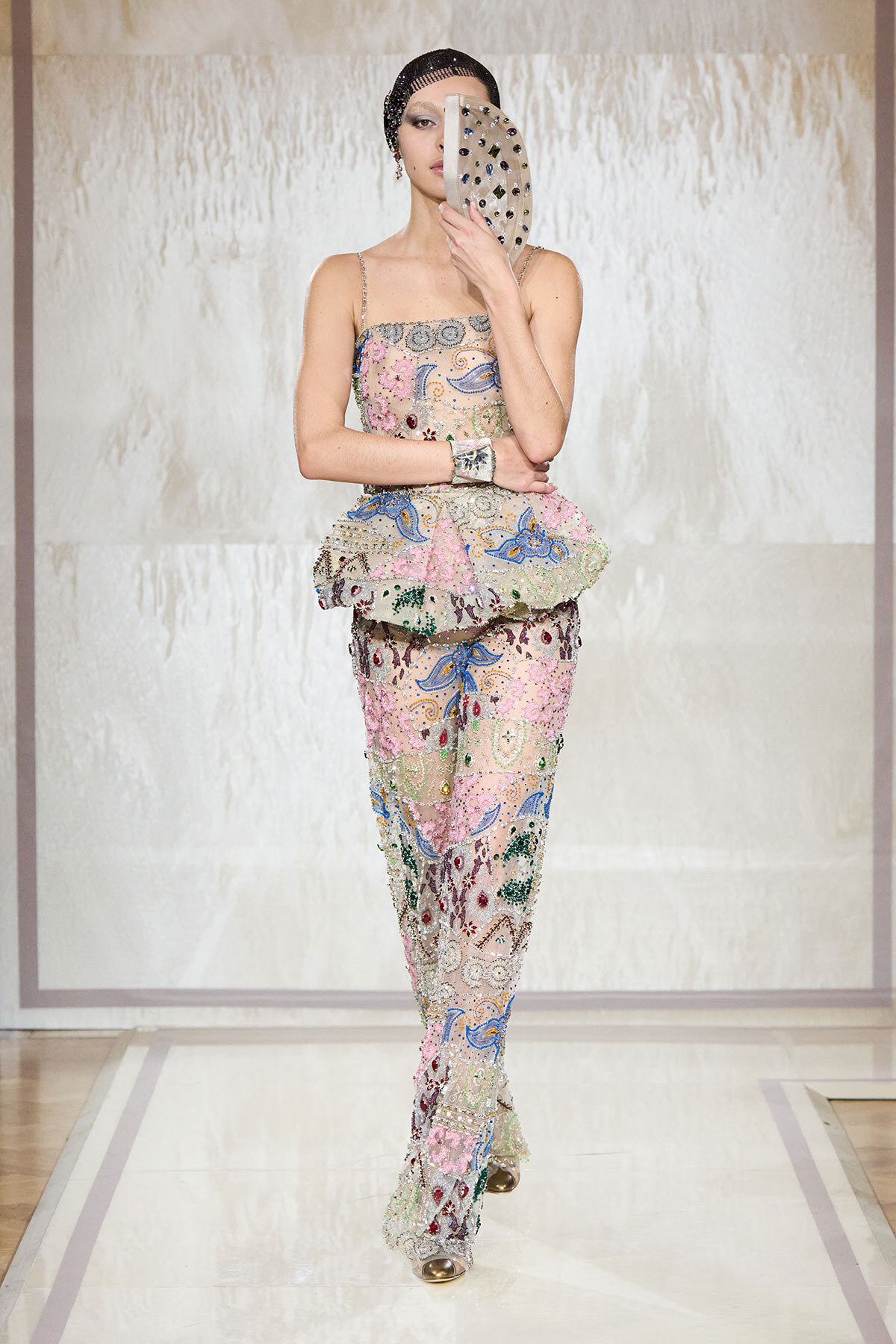 Model walking a runway in a pastel embroidered jumpsuit with floral and peacock motifs, holding a beaded circular fan over her face.