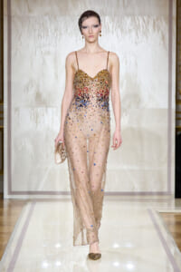 Model walks the runway in a sheer nude gown adorned with multicolor crystals and gold detailing, holding a small metallic clutch.