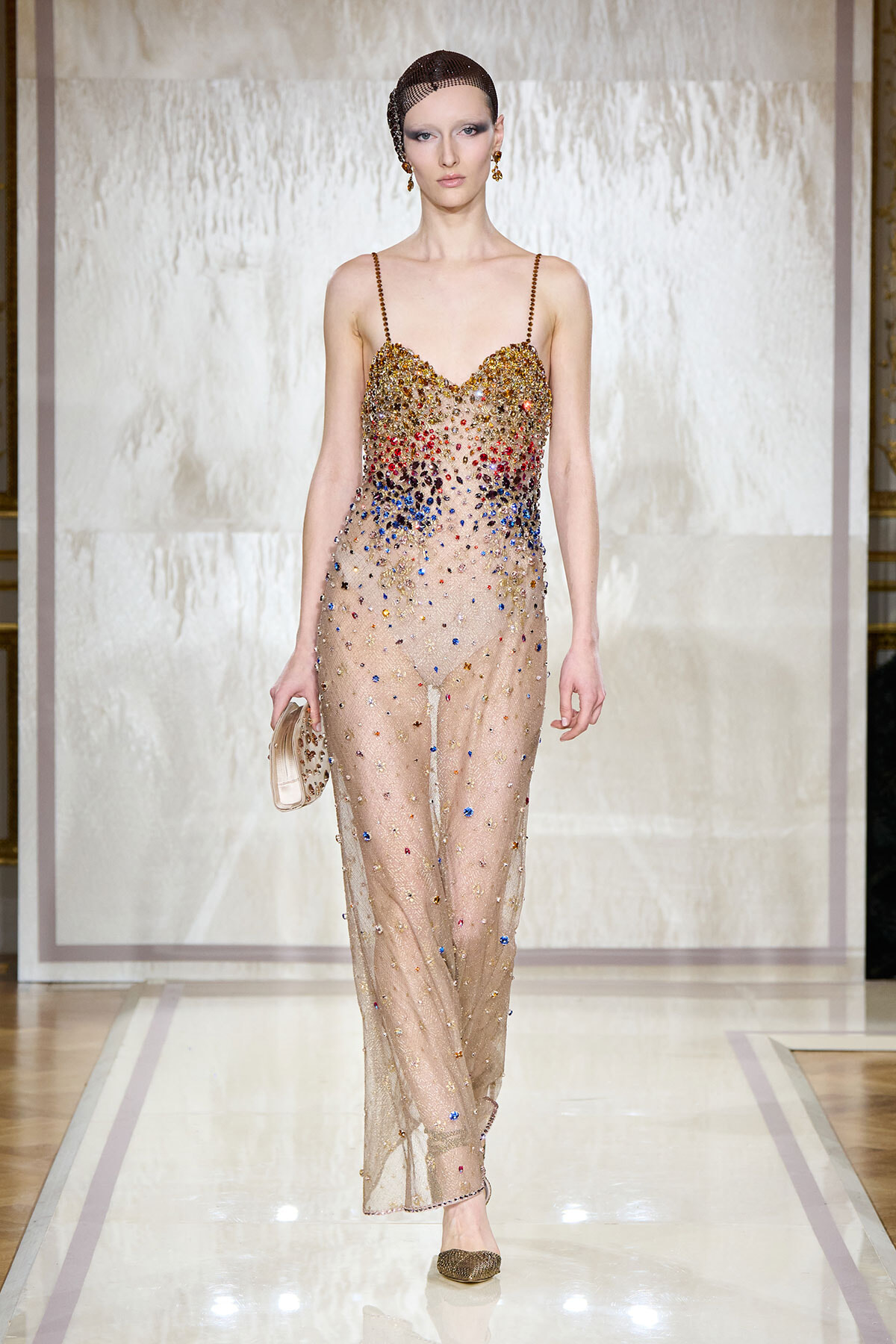 Model walks the runway in a sheer nude gown adorned with multicolor crystals and gold detailing, holding a small metallic clutch.