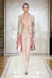 Model walks the runway in a champagne beaded sheer jumpsuit and metallic floral-printed coat.