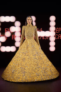 Model in a full-length gold sequined gown walking a runway, with pink bokeh lights in the background.