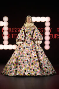 Model wearing a colorful patchwork quilted ball gown with enormous puff sleeves and a ruffled high collar on a runway stage.