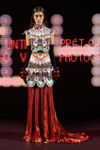 Model on a runway wearing an ornate, mosaic-patterned top with a high collar and floral details, paired with long red satin fringe skirt and matching sleeves/armwear.
