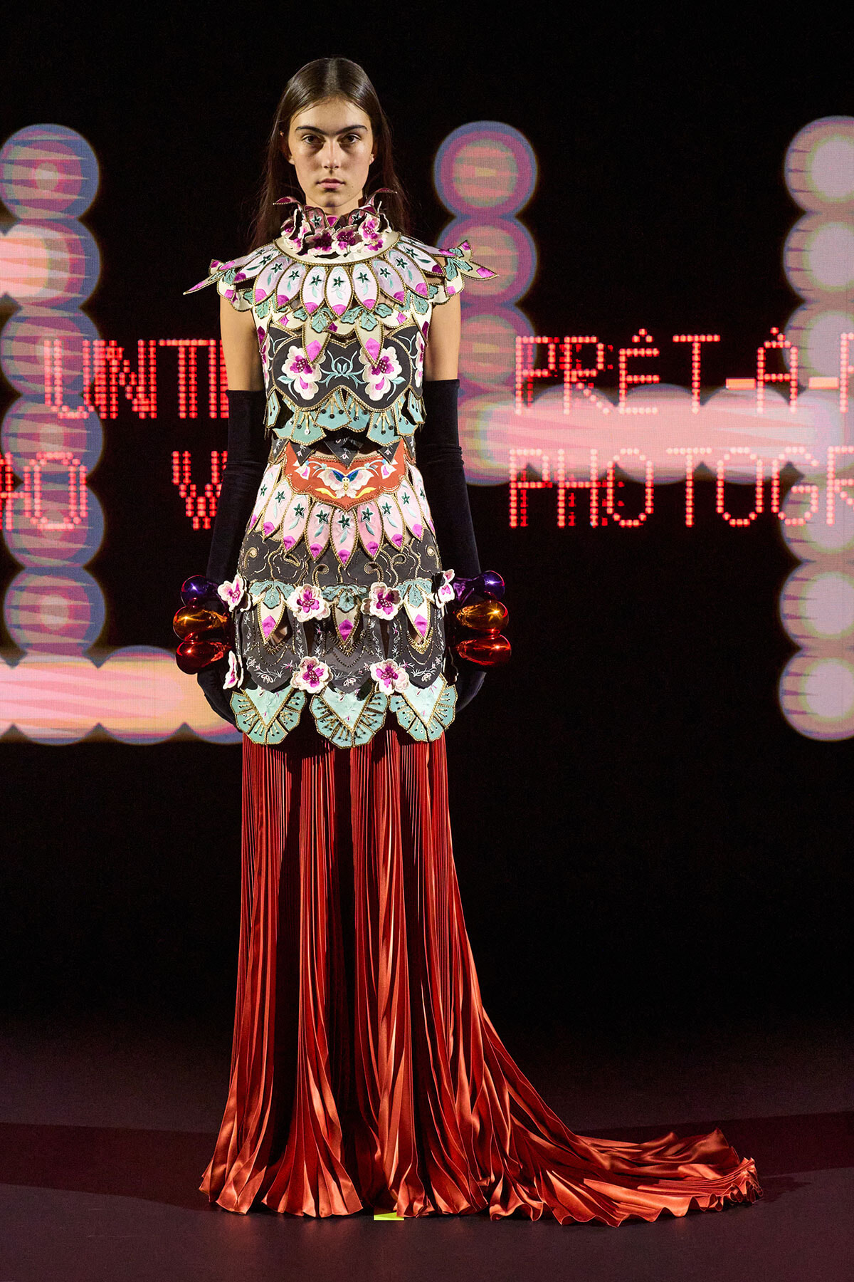 Model on a runway wearing an ornate, mosaic-patterned top with a high collar and floral details, paired with long red satin fringe skirt and matching sleeves/armwear.