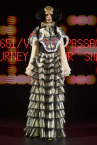 Model on a runway wearing a metallic silver, multi-layered ruffled gown with butterfly embroidery and large black headpieces against a dark background with red lights.