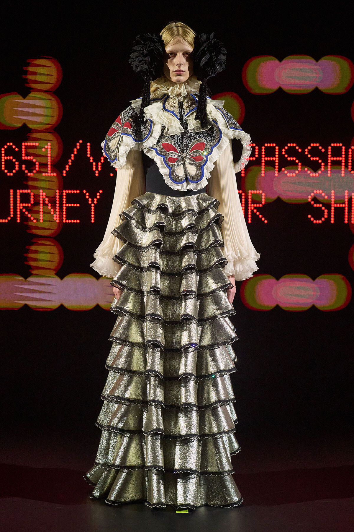 Model on a runway wearing a metallic silver, multi-layered ruffled gown with butterfly embroidery and large black headpieces against a dark background with red lights.
