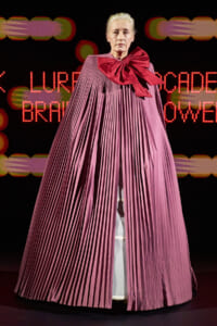 Model on a runway wearing a long, pleated pink cape with a large red bow at the neck, neon backdrop in the background.