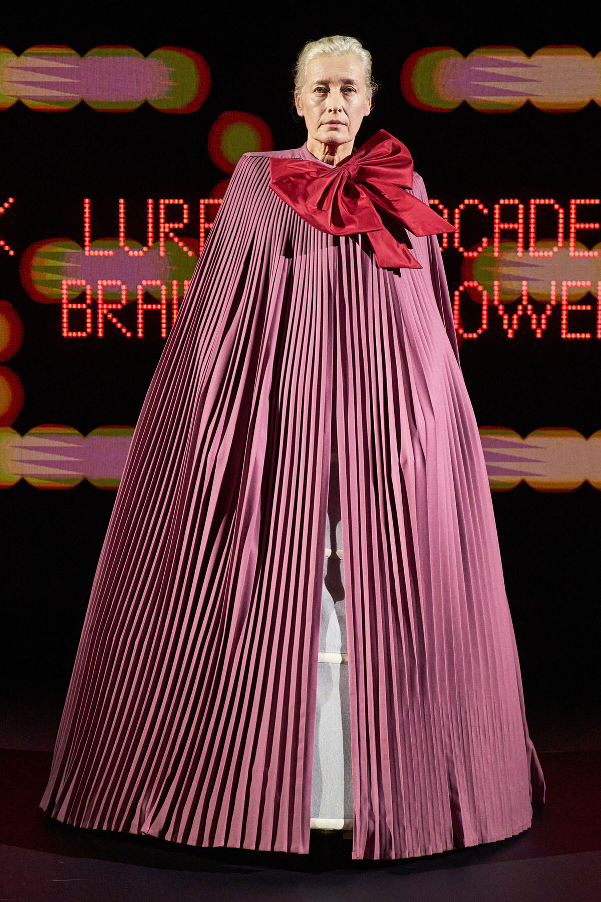 Model on a runway wearing a long, pleated pink cape with a large red bow at the neck, neon backdrop in the background.