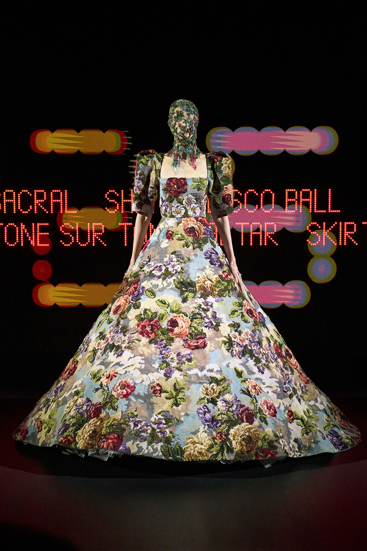 Model in a full, floral ball gown with puff sleeves, standing against a black backdrop with red neon text behind.