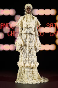 Model on runway wearing an ivory lace gown with layered ruffles and a lace headpiece, standing against a blurred lights backdrop.