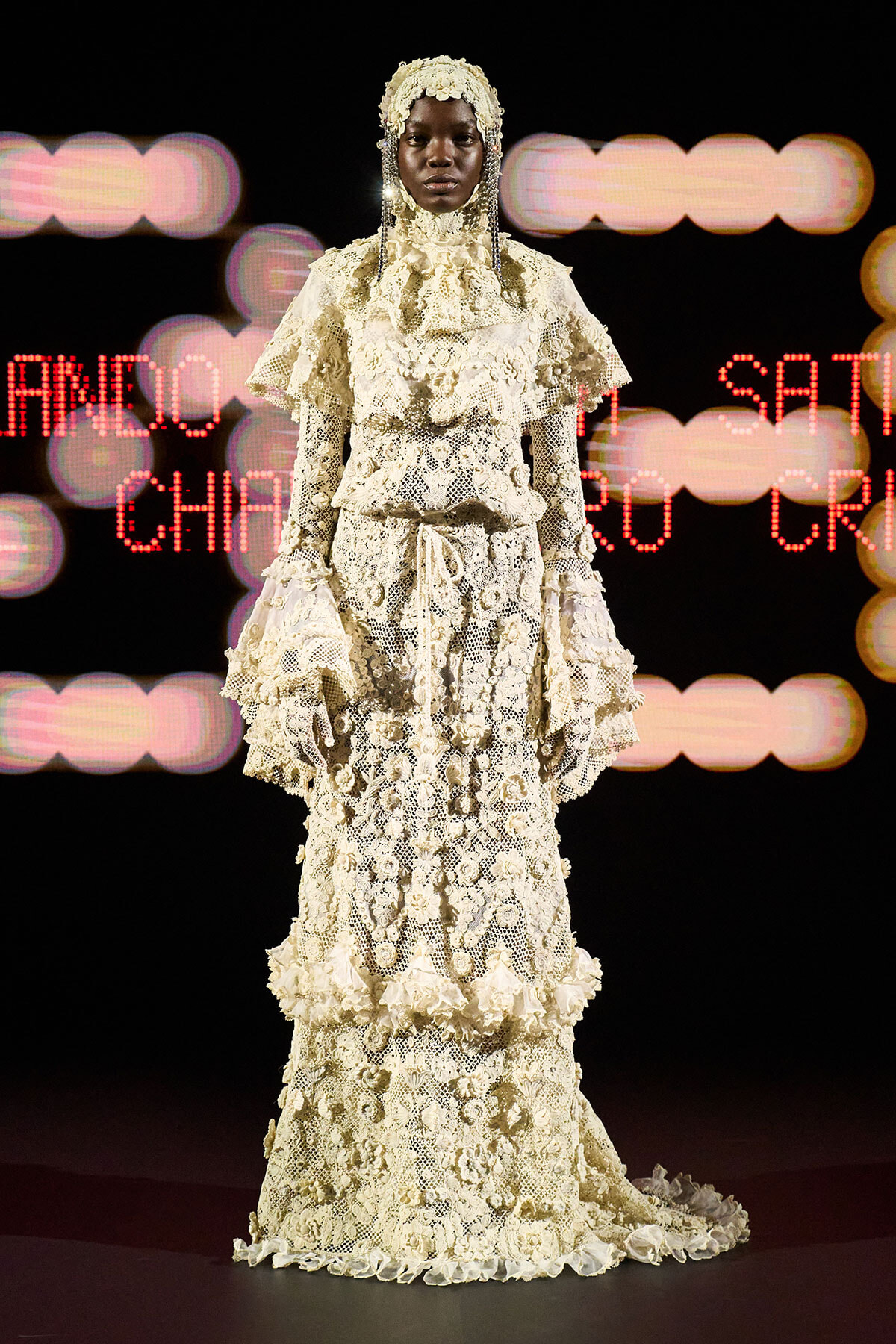 Model on runway wearing an ivory lace gown with layered ruffles and a lace headpiece, standing against a blurred lights backdrop.