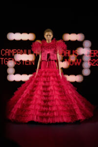Model wearing a dramatic bright red ruffled ballgown on a runway, backdrop of pink LED text and lights.
