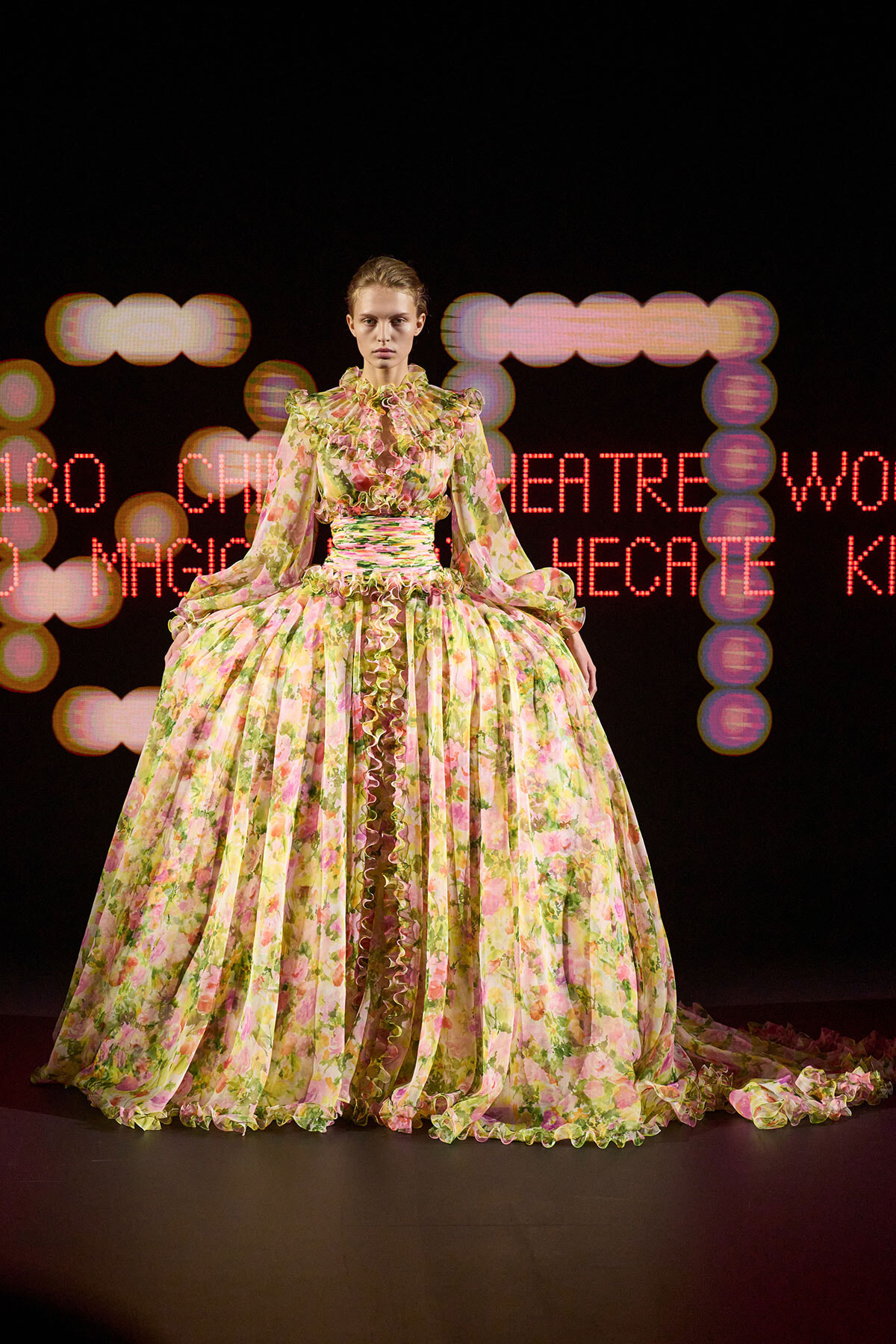 Model wearing a voluminous pastel floral ball gown on a fashion-show runway, with a ruffled high collar and long sleeves.