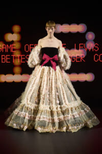 Model wearing a voluminous ivory lace gown with pink floral trim and a large pink bow at the waist, posing on a runway.