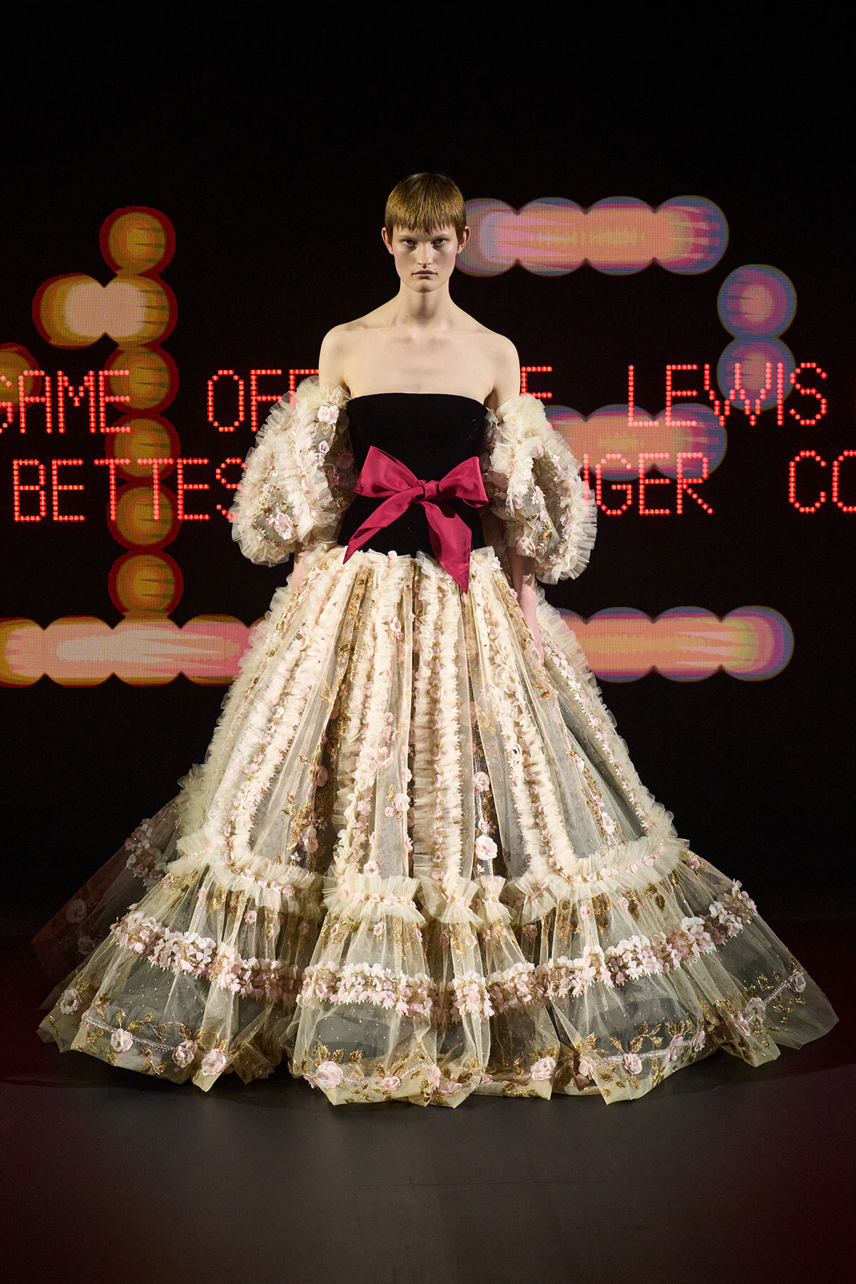 Model wearing a voluminous ivory lace gown with pink floral trim and a large pink bow at the waist, posing on a runway.