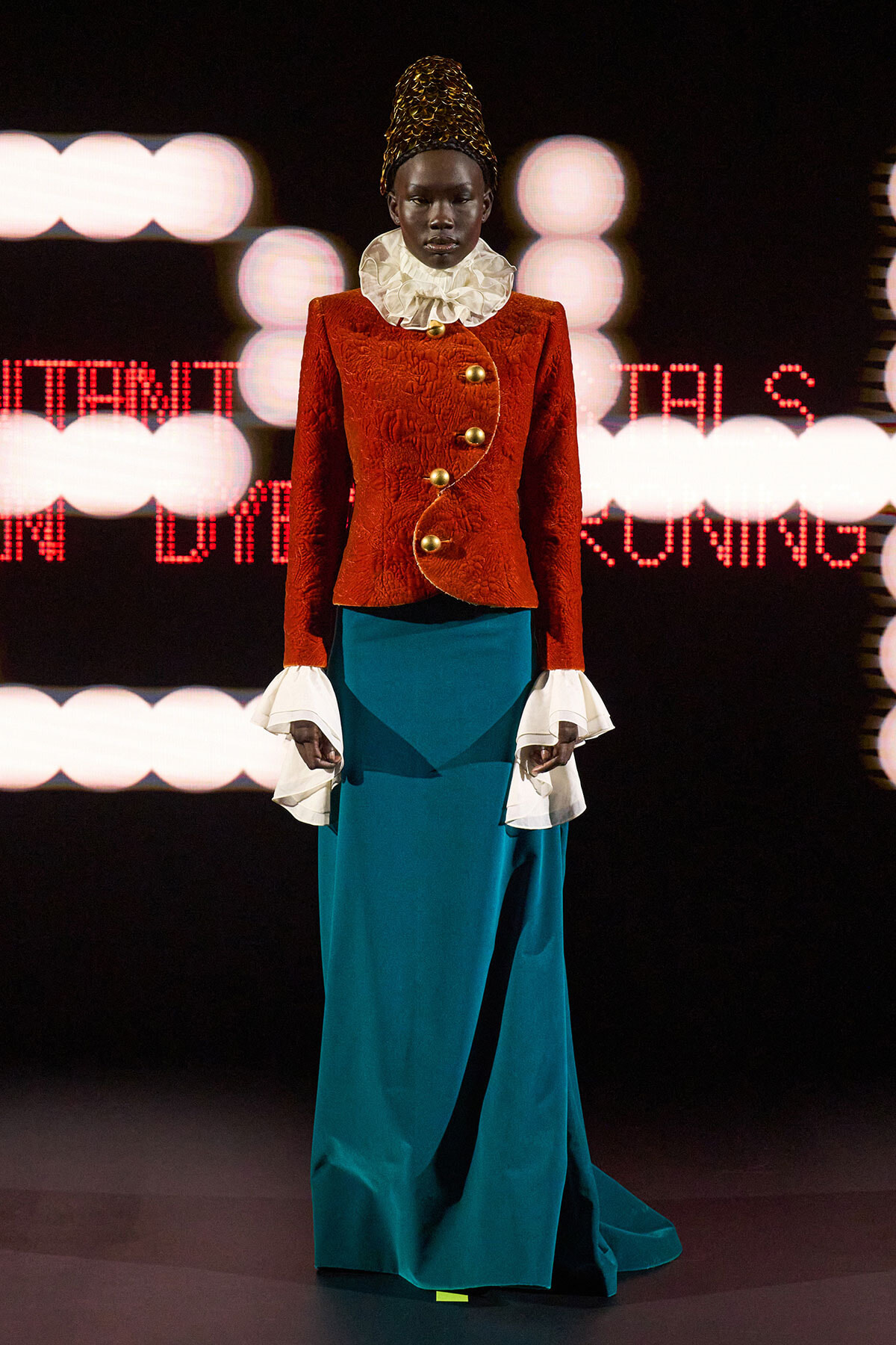 Model on runway wearing a red textured jacket with gold buttons, a white ruffled collar, and a teal skirt.