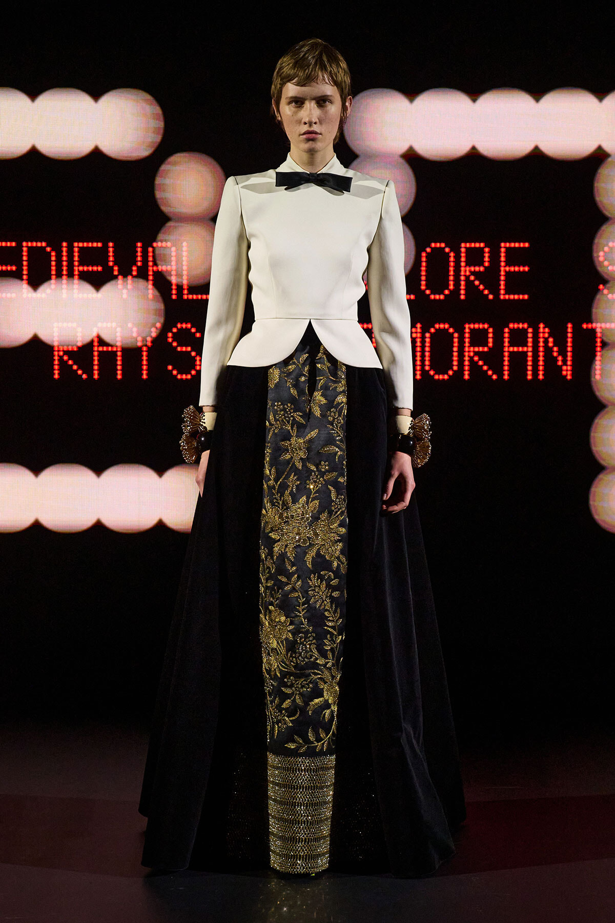 Model on runway wearing a white structured jacket with a black bow tie and a long black skirt with gold floral embroidery; dramatic stage lights in background.