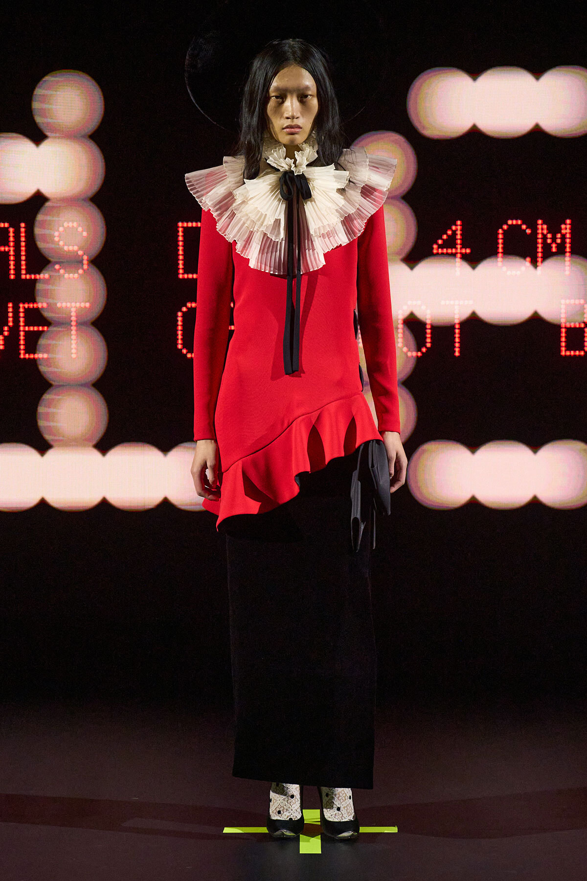 Runway model wearing a red dress with an oversized white ruffled collar and black bow, paired with a black skirt on a lit stage.