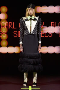 Runway model in a polka-dot blazer with large white lapels, lime bow, and a voluminous multi-ring black skirt, wearing lace gloves and glitter boots.