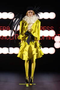 Runway model wearing a neon yellow, layered ruffle coat with a white feather collar, black bow belt, matching yellow lace tights, and a black feathered hat perched on the head.