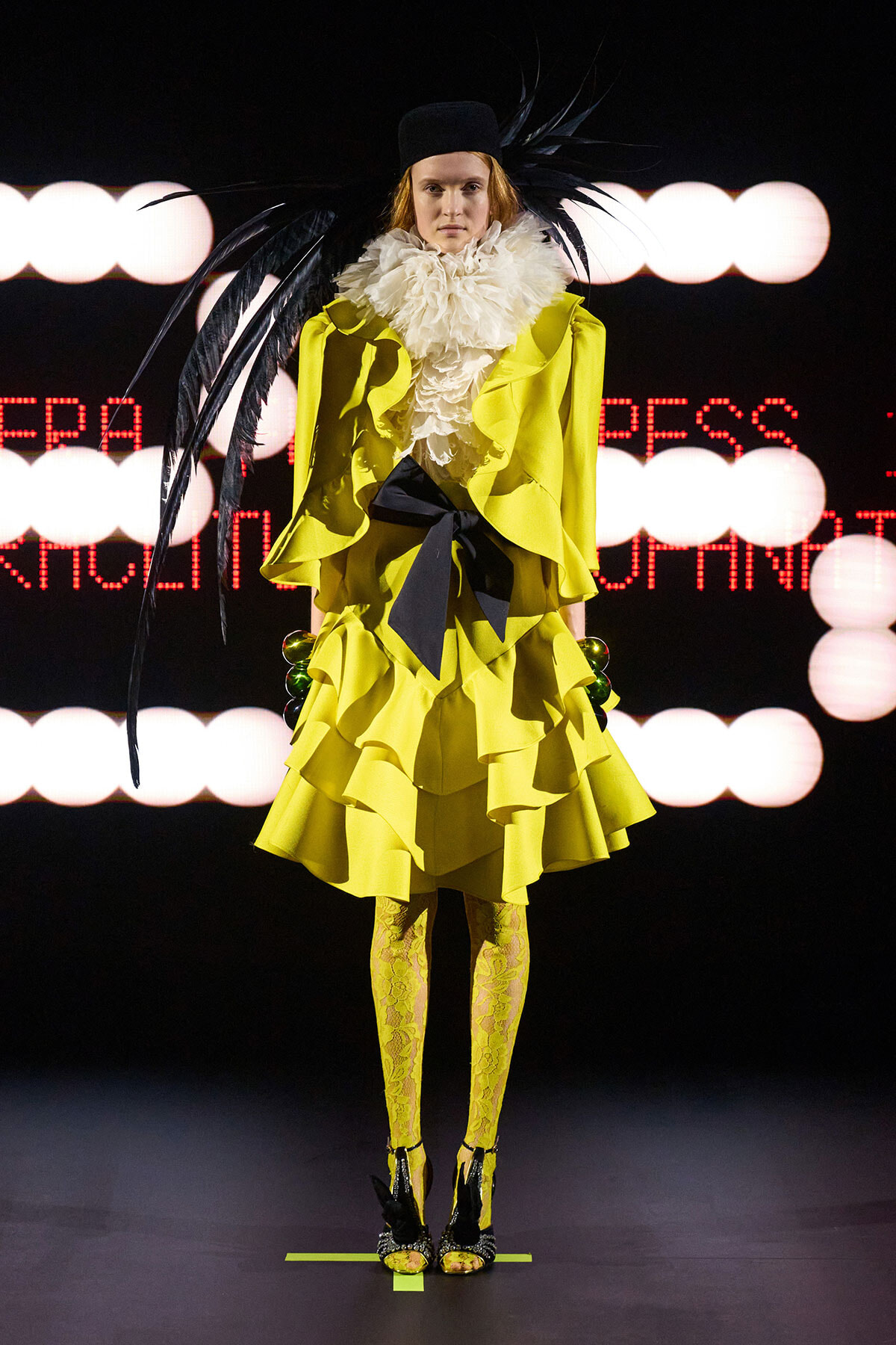 Runway model wearing a neon yellow, layered ruffle coat with a white feather collar, black bow belt, matching yellow lace tights, and a black feathered hat perched on the head.