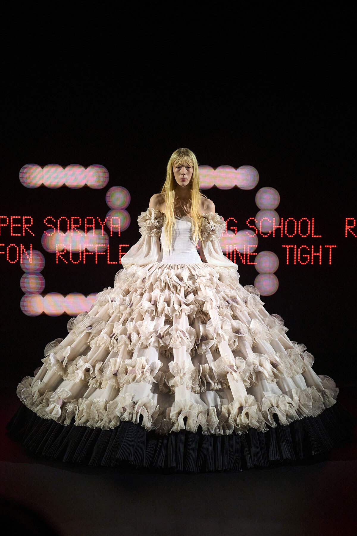 Model in a voluminous white ruffled ball gown stands on a dark runway, neon red text behind her on stage.