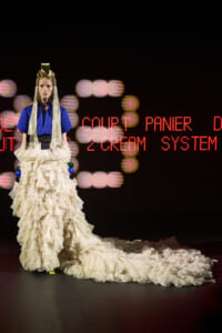 Model on a runway wearing a cobalt blue top and a dramatic layered ivory gown with a long train and a gold headpiece, against a neon sign backdrop.