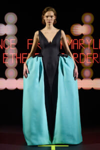 Model on runway wearing a black V-neck dress with a teal, cape-like over-skirt that opens at the center front.
