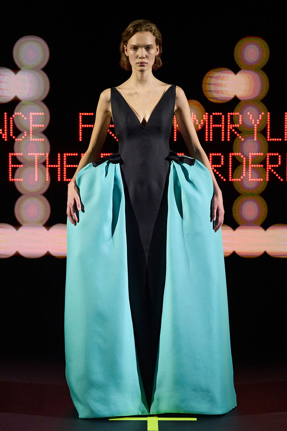 Model on runway wearing a black V-neck dress with a teal, cape-like over-skirt that opens at the center front.