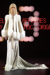 Model in a white gown with voluminous fur trim at shoulders and cuffs, standing on a runway with a long train and blurred neon signage behind.