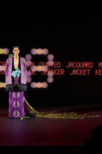 Model wearing a purple embroidered robe with teal sash and gold train on a runway with red text backdrop.