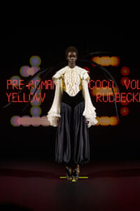 Model wearing a cream ruffled blouse with gold accents and a black corset over wide, dark pants on a runway, with neon red text and colorful lights in the background.