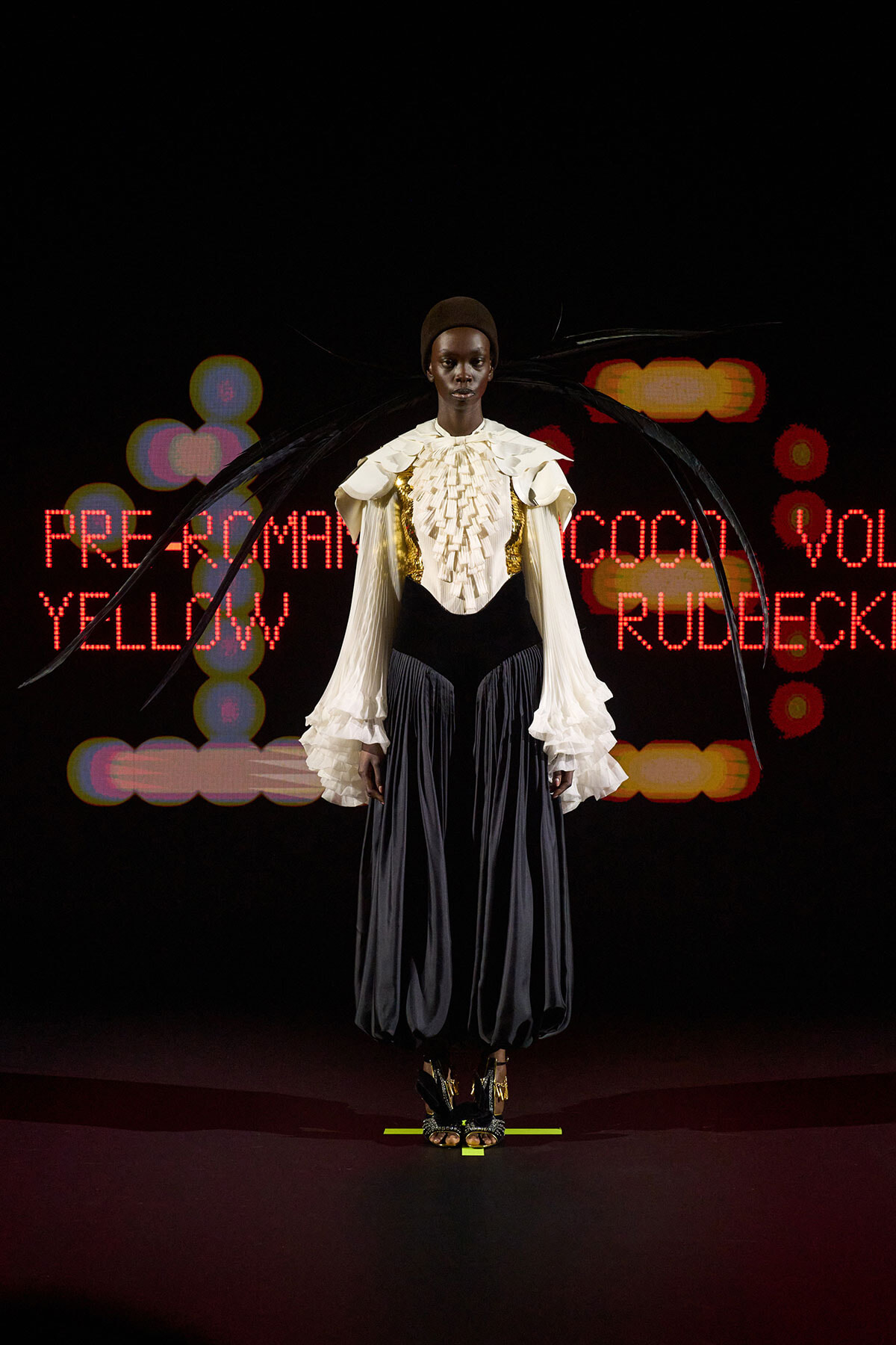 Model wearing a cream ruffled blouse with gold accents and a black corset over wide, dark pants on a runway, with neon red text and colorful lights in the background.