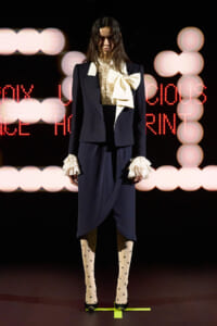 Model on runway wearing a navy tailored suit with a cream ruffled blouse and oversized bow, plus ruffled cuffs and polka-dot tights.