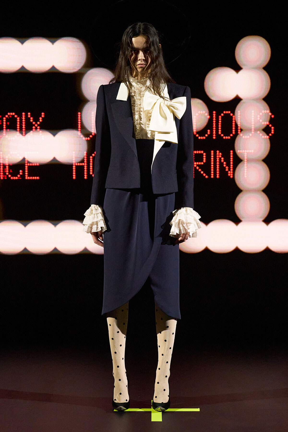 Model on runway wearing a navy tailored suit with a cream ruffled blouse and oversized bow, plus ruffled cuffs and polka-dot tights.