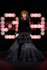 Model wearing a dramatic black Victorian-style gown with tiered, pleated skirts and ruffled cuffs on a runway, with a red neck scarf and pink bokeh lights in the background.