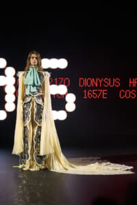 Model on a runway wearing an ornate cream and silver gown with a teal neck bow and flowing cape, against a backdrop of red LED text and lights.