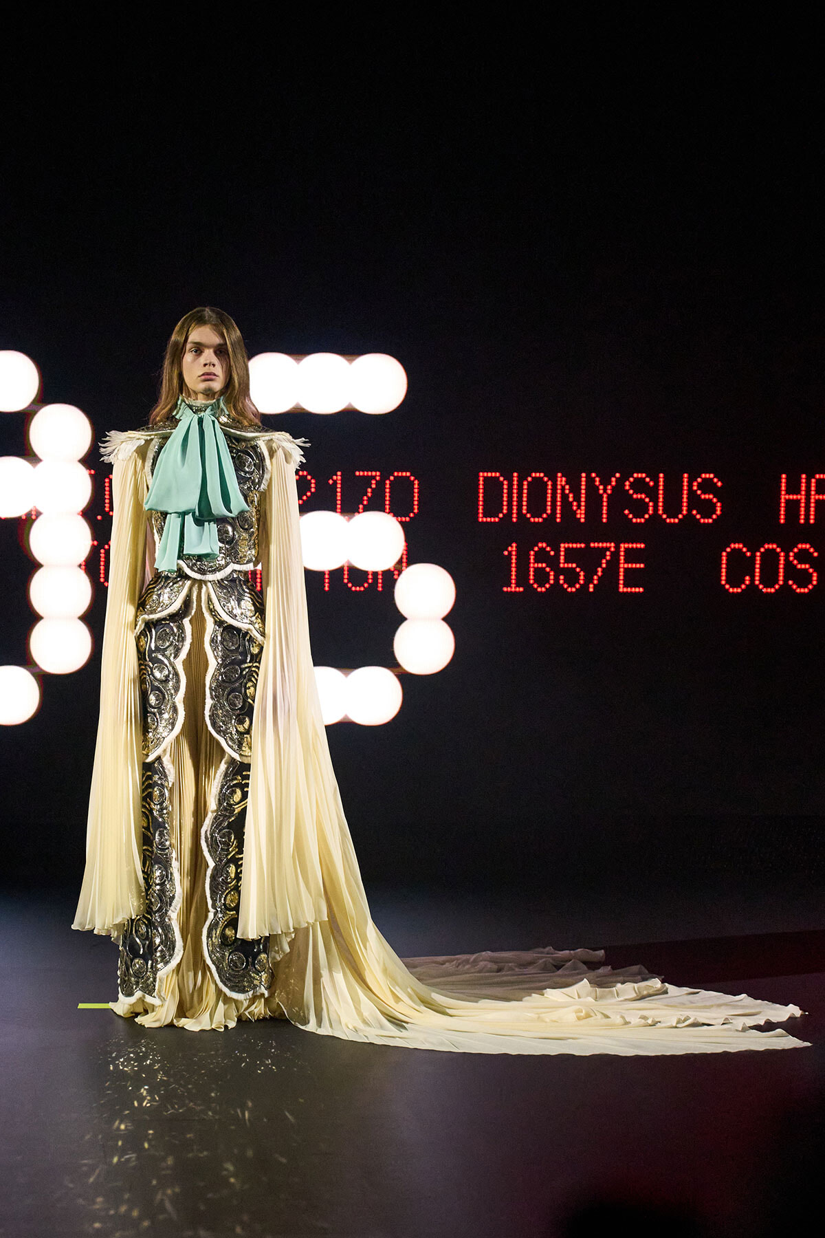 Model on a runway wearing an ornate cream and silver gown with a teal neck bow and flowing cape, against a backdrop of red LED text and lights.