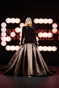 Model on a runway wearing a black long-sleeve top and a voluminous gray satin ball gown, with circular stage lights and red LED text behind.