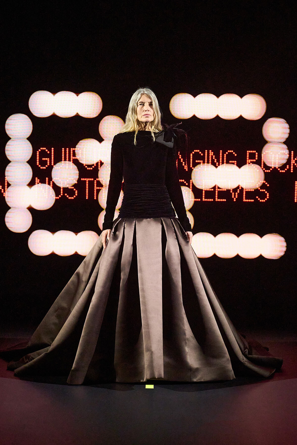 Model on a runway wearing a black long-sleeve top and a voluminous gray satin ball gown, with circular stage lights and red LED text behind.