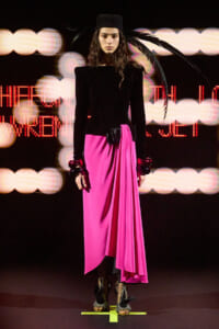 Model on a runway wearing a black velvet top, bright pink draped skirt, black belt, feathered accessory, and strappy heels against a neon-lit background.