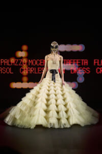 Model on a dark runway wearing a strapless pale yellow tiered ball gown and an ornate jeweled mask.