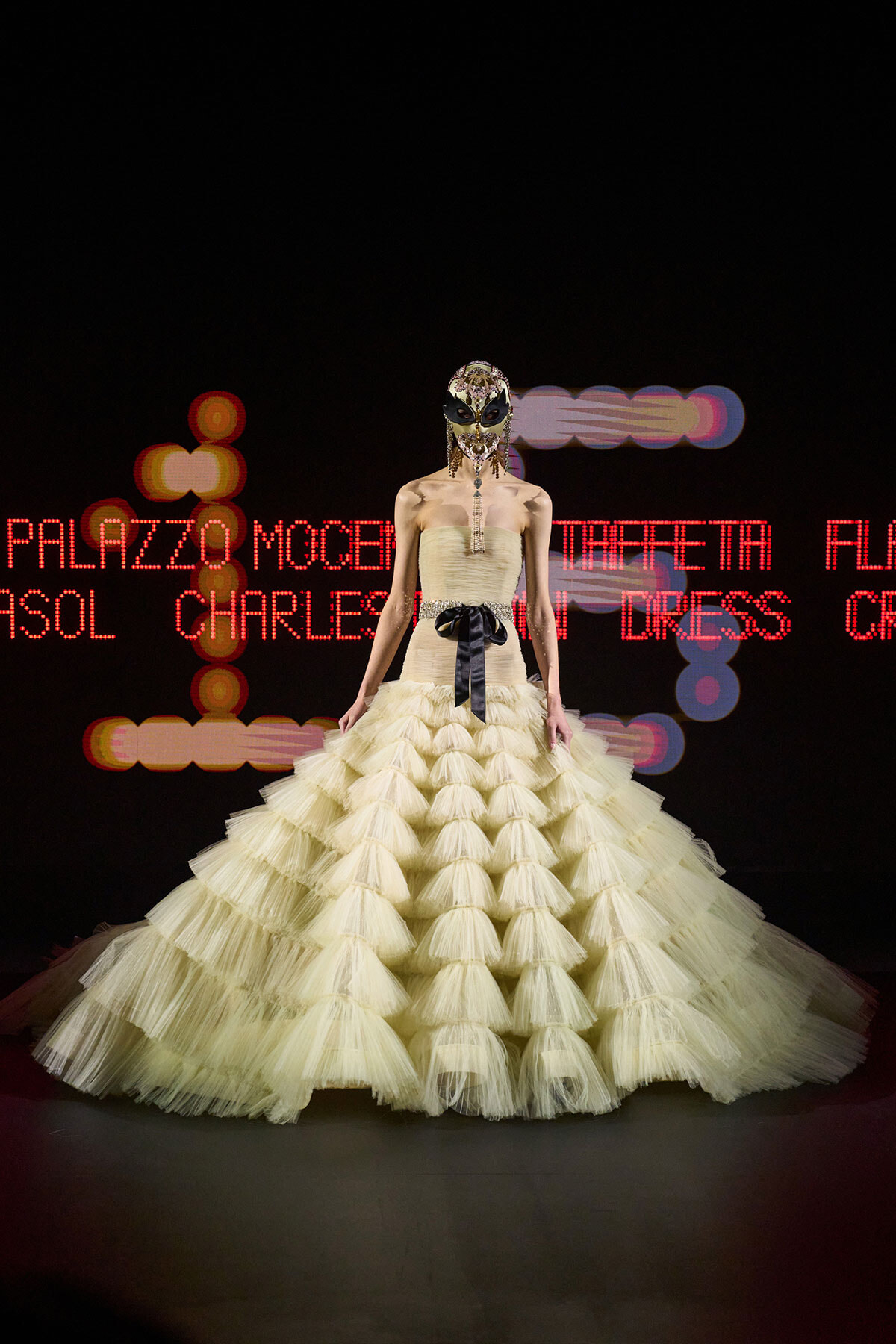 Model on a dark runway wearing a strapless pale yellow tiered ball gown and an ornate jeweled mask.