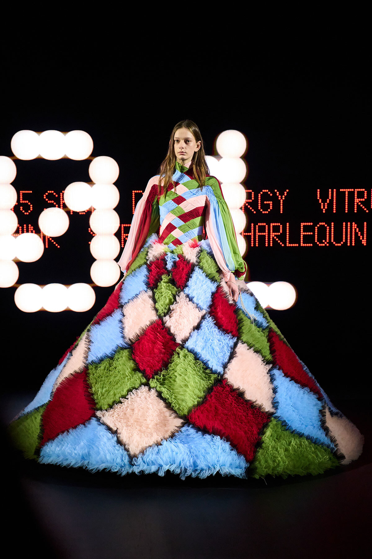 Model wearing a voluminous patchwork gown of diamond patches in pink, red, blue, green on a dark runway with bright circular lights in the background.