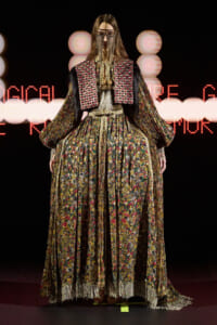 Model on a runway wearing an ornate, multicolored floral gown with gold fringe and beaded face veil.